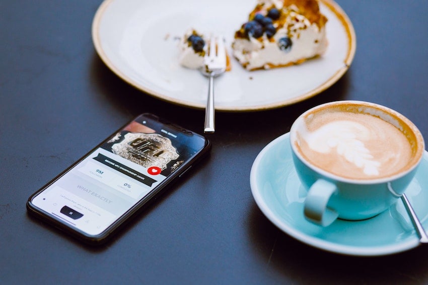 A smartphone next to a cup of coffee and a piece of cheesecake.